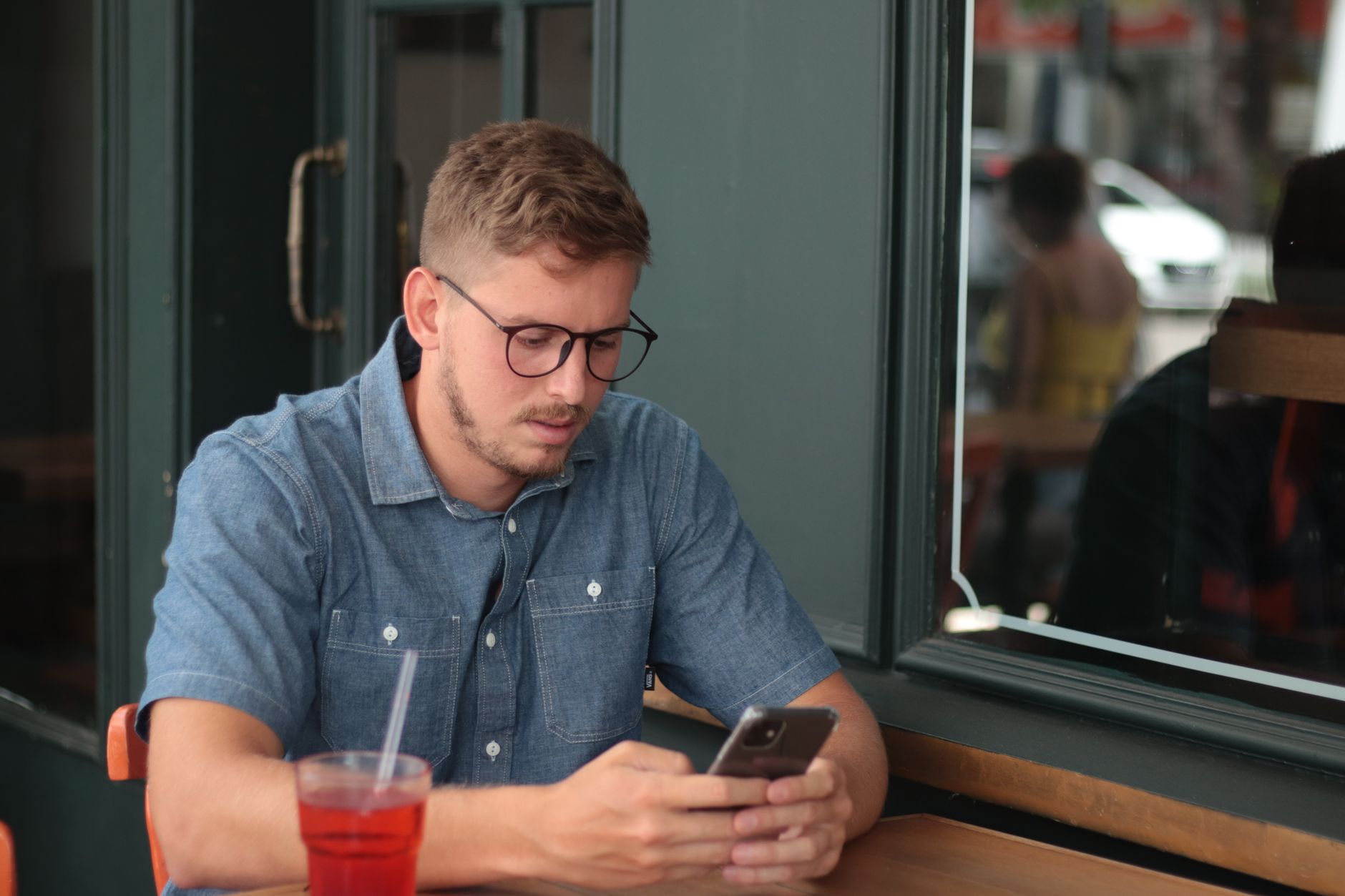 A young man in round glasses and a denim shirt reading his phone at a café table beside a red drink