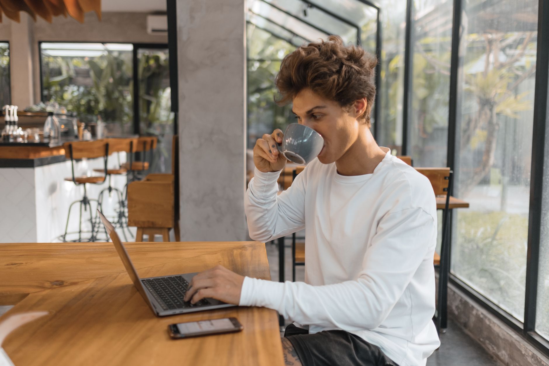 A young man with tousled hair sipping coffee while typing on a laptop at a wooden café table